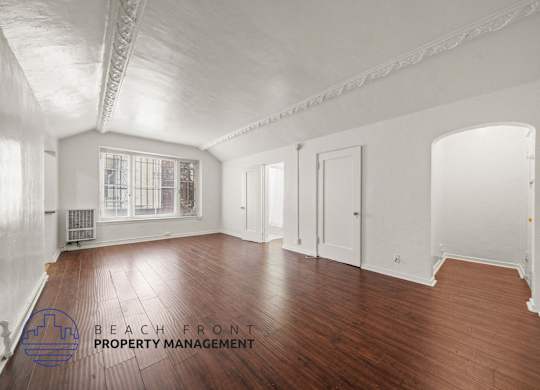 A spacious room with wooden floors and white walls, with a window and a door visible.