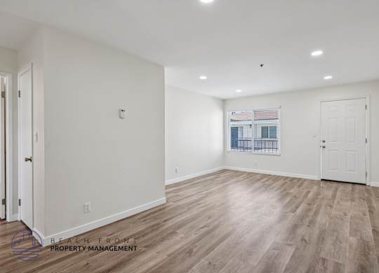 A spacious room with wooden flooring and white walls, with a window showing a view of a balcony.