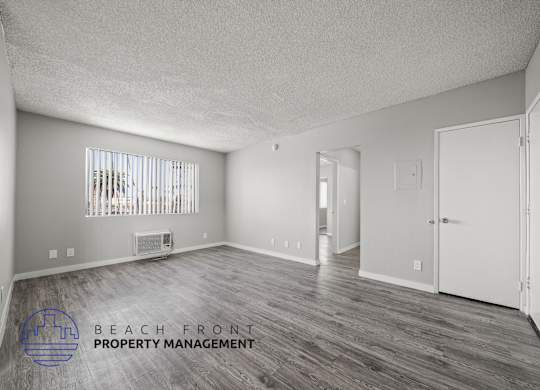 A spacious room with wooden flooring and white walls, with a window covered by vertical blinds.