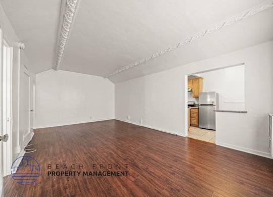 A spacious room with wooden floors and white walls, with a kitchenette visible in the background.