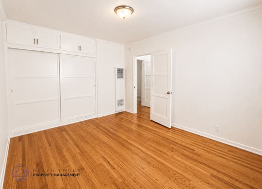 A spacious room with wooden floors and white walls, featuring a set of white built-in cabinets and a doorway leading to another room.