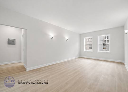 A spacious room with wooden flooring and white walls, featuring a window with blinds and a wall-mounted light fixture.