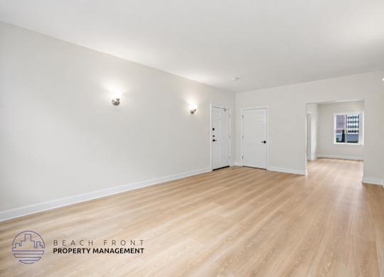 A spacious room with wooden floors and white walls, with a logo for Beach Front Property Management in the bottom left corner.