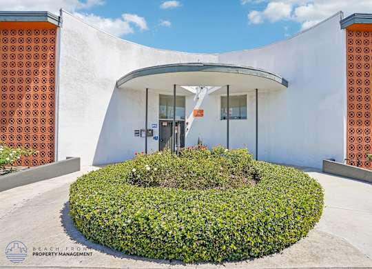 A white building with orange patterned walls and a circular green bush in front.
