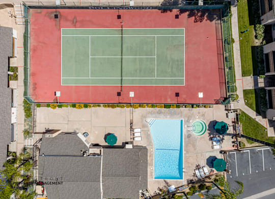 A tennis court is surrounded by a pool and a parking lot.