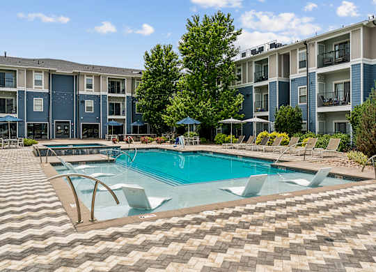 A swimming pool surrounded by a tiled patio and lounge chairs in front of apartment buildings.