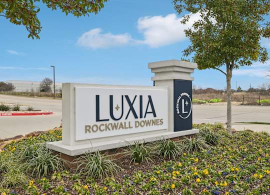 A sign for Luxia Rockwall Downes in front of a tree and a fence.