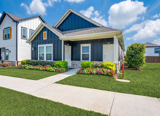 A house with a blue roof and white walls is surrounded by a well-manicured lawn and colorful flower beds.