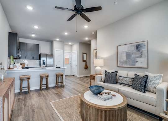 A modern living room with a ceiling fan and a kitchen area in the background.