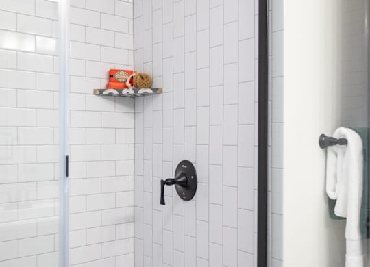 A white tiled shower with a black faucet and a black and white towel hanging on the door.