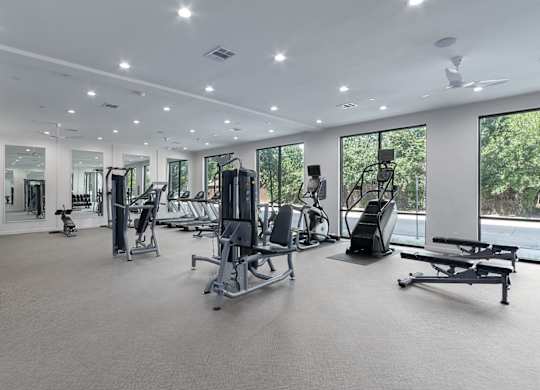 A spacious gym with a variety of equipment and a view of trees outside the windows.