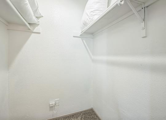 A white wall with a small electrical outlet and a white shelf above a carpeted floor.