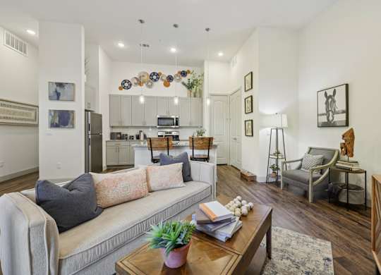 An apartment living room featuring a plush beige sofa and warm decor, looking back toward the bright kitchen with its dark wood floors.