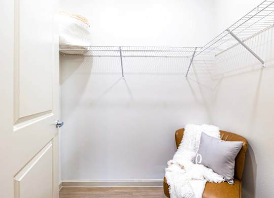 A white chair with a grey pillow and a white blanket is in front of a white wall with a metal shelf above it.