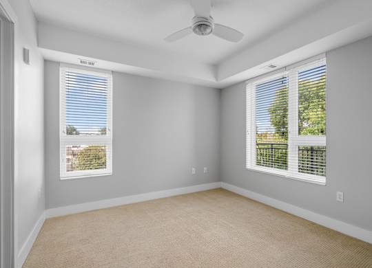 A room with a fan, two windows, and a carpeted floor.