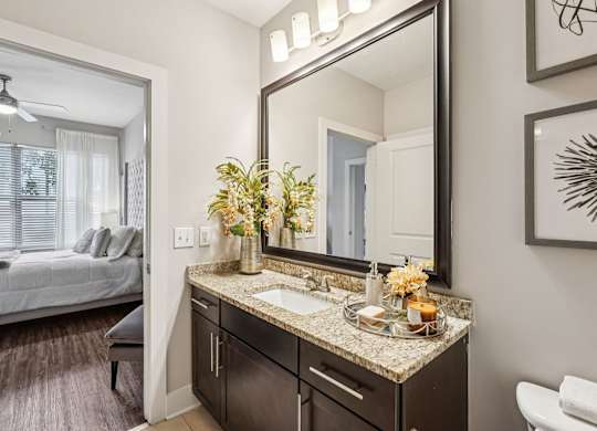 A bathroom with a large mirror, a sink, and a bedroom with a bed and a window.