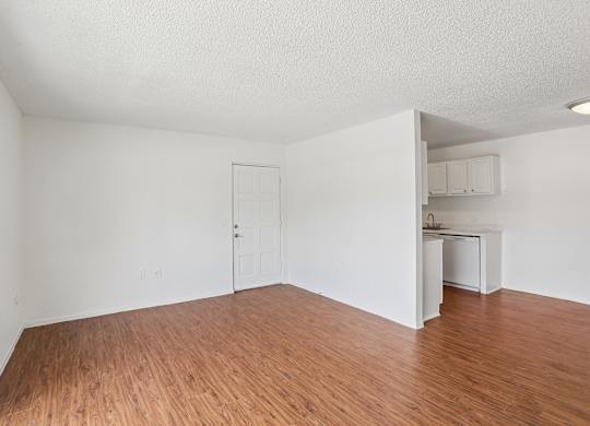 A room with wooden floors and white walls, with a kitchenette visible in the background.