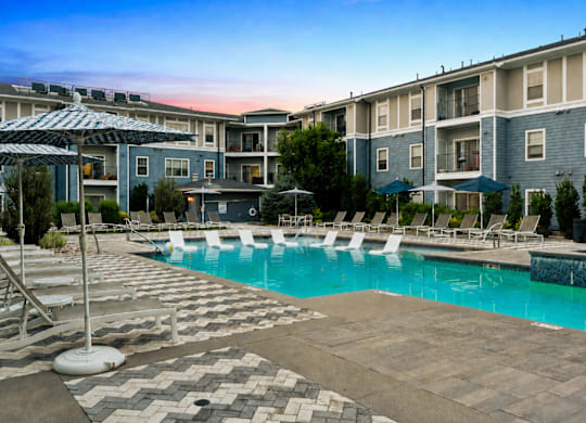 A swimming pool surrounded by lounge chairs and umbrellas in front of apartment buildings.