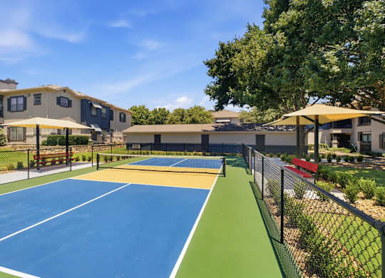 A tennis court is surrounded by a fence and there are two buildings in the background.