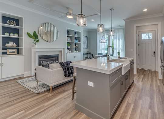A modern builder grade kitchen with a grey cabinets, wooden floor and front entryway and living room with fireplace in the background.