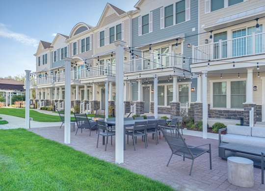 A townhome complex patio with fire pit, table, and chairs on a brick walkway underneath cafe lights.