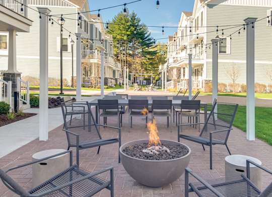 A townhome complex patio with fire pit, table and chairs on a brick walkway underneath cafe lights.