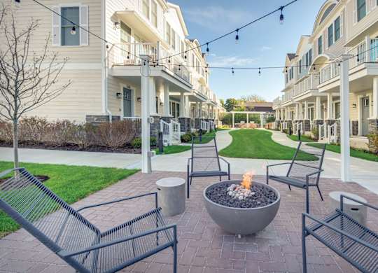 A townhome complex patio with fire pit and chairs on a brick walkway underneath cafe lights.