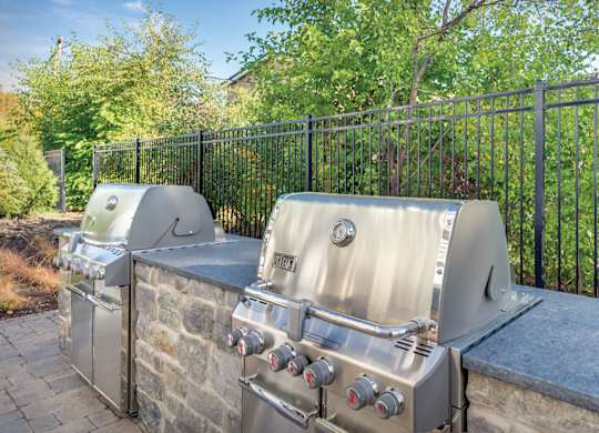 A silver grill is on a stone slab with a black fence and trees in the background.