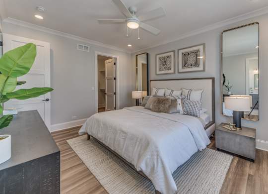 A modern builder grade bedroom with a large bed, a mirror, ceiling fan, and walk in closet.