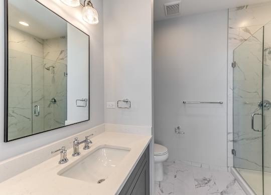 A modern builder grade bathroom with vanity, mirror, toilet, and glass tiled shower.