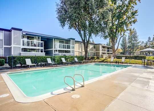 A swimming pool surrounded by trees and chairs in front of apartment buildings.