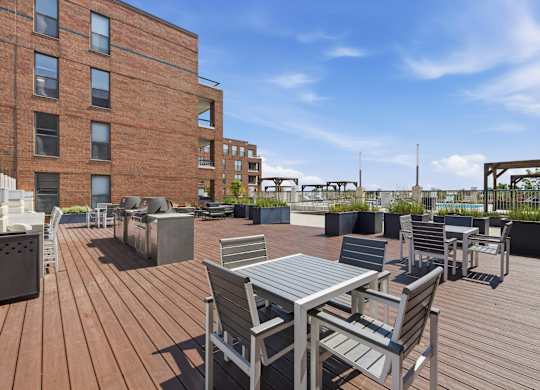 A rooftop wooden deck with metal tables and chairs in front of a grilling statioin.