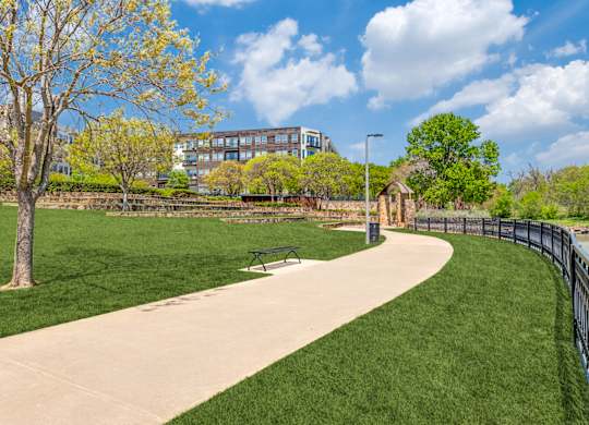 A park with a paved walkway, a bench, and a fence.
