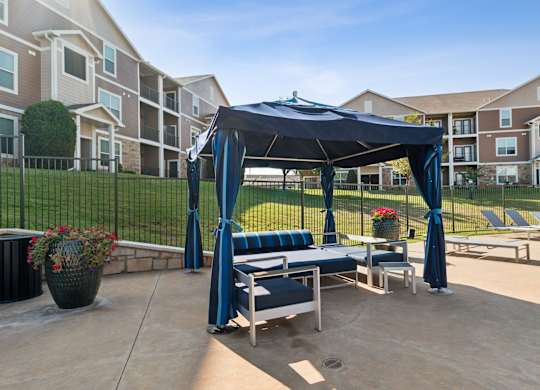 A gazebo with a blue canopy is situated in a courtyard with lounge chairs and potted plants.
