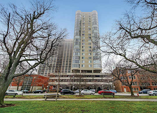 A tall building with many windows is surrounded by trees and cars.