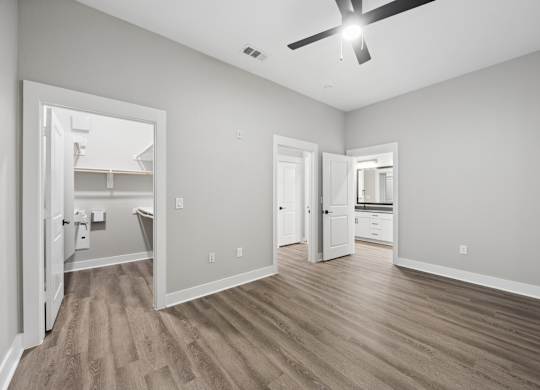 A spacious room with a ceiling fan and light, hardwood floors, and a view into another room through an open door.