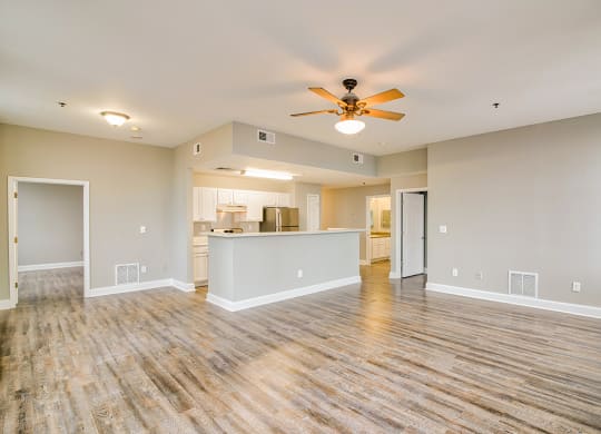 an empty living room with a ceiling fan and a kitchen in the background
