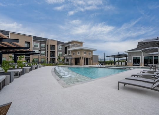 A swimming pool surrounded by sun loungers and umbrellas in front of apartment buildings.