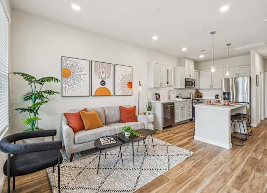 A modern kitchen and living room with a grey couch and orange pillows.