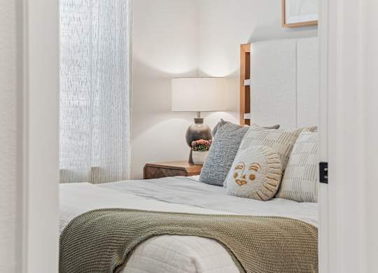 A bedroom with a bed, a nightstand with a lamp, and a window with curtains.