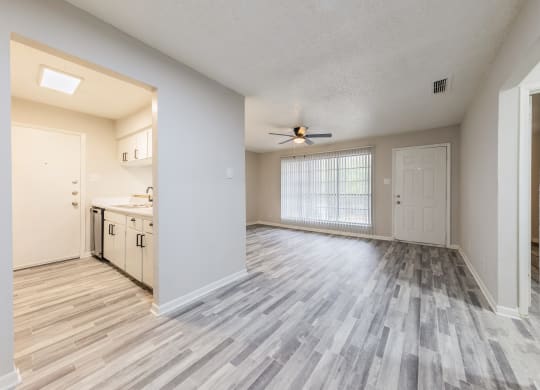an empty living room with a ceiling fan and a kitchen in the background