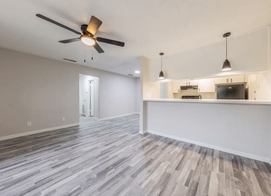 an empty living room with a ceiling fan and a kitchen in the background