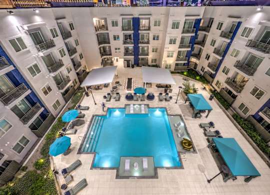 A large swimming pool surrounded by lounge chairs and umbrellas in a courtyard surrounded by apartment buildings.