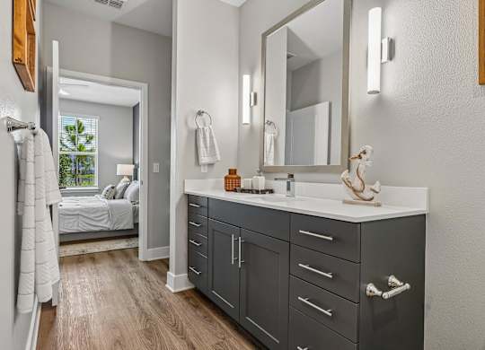 A bathroom with a vanity, mirror, and a bedroom in the background.