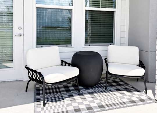A black and white checkered rug sits on a patio with two chairs and a round ottoman.