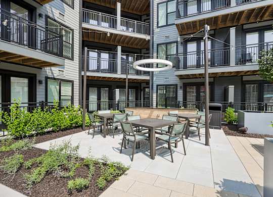 A patio with a table and chairs is surrounded by plants and a building with balconies.