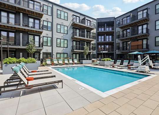 A swimming pool surrounded by lounge chairs and umbrellas in front of apartment buildings.