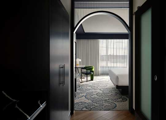 A room with a bed, a chair and a table is visible through an arched doorway.