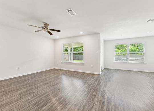 Empty room with gray wood-look flooring, white walls, ceiling fan, and two windows with blinds.