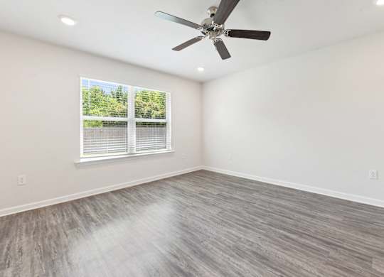Empty room with gray wood flooring, white walls, ceiling fan, and window.